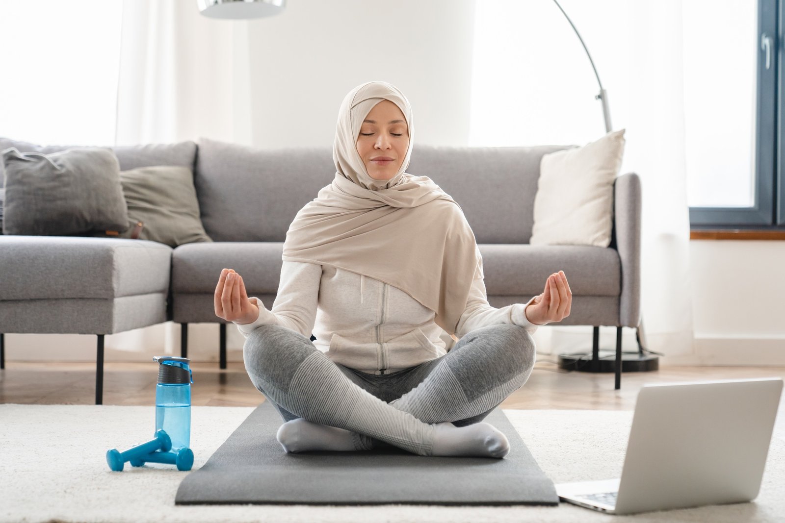 Arabian muslim islamic woman in hijab meditating on exercise mat in the living room using laptop