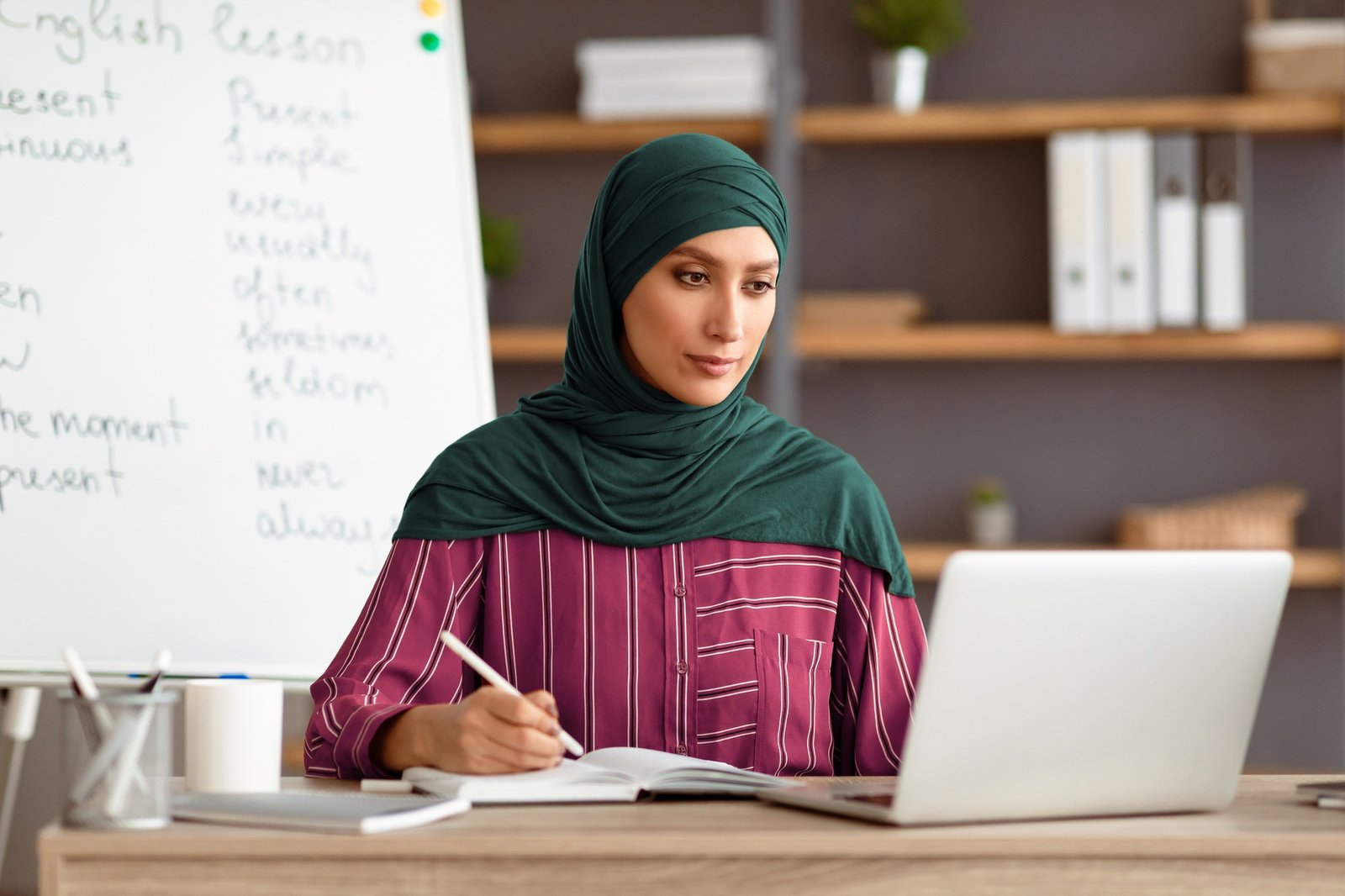Islamic teacher in headscarf sitting at desk looking at laptop